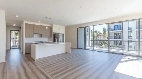 An empty open-plan apartment with a bare kitchen island, wood-look floors and floor-to-ceiling balcony doors